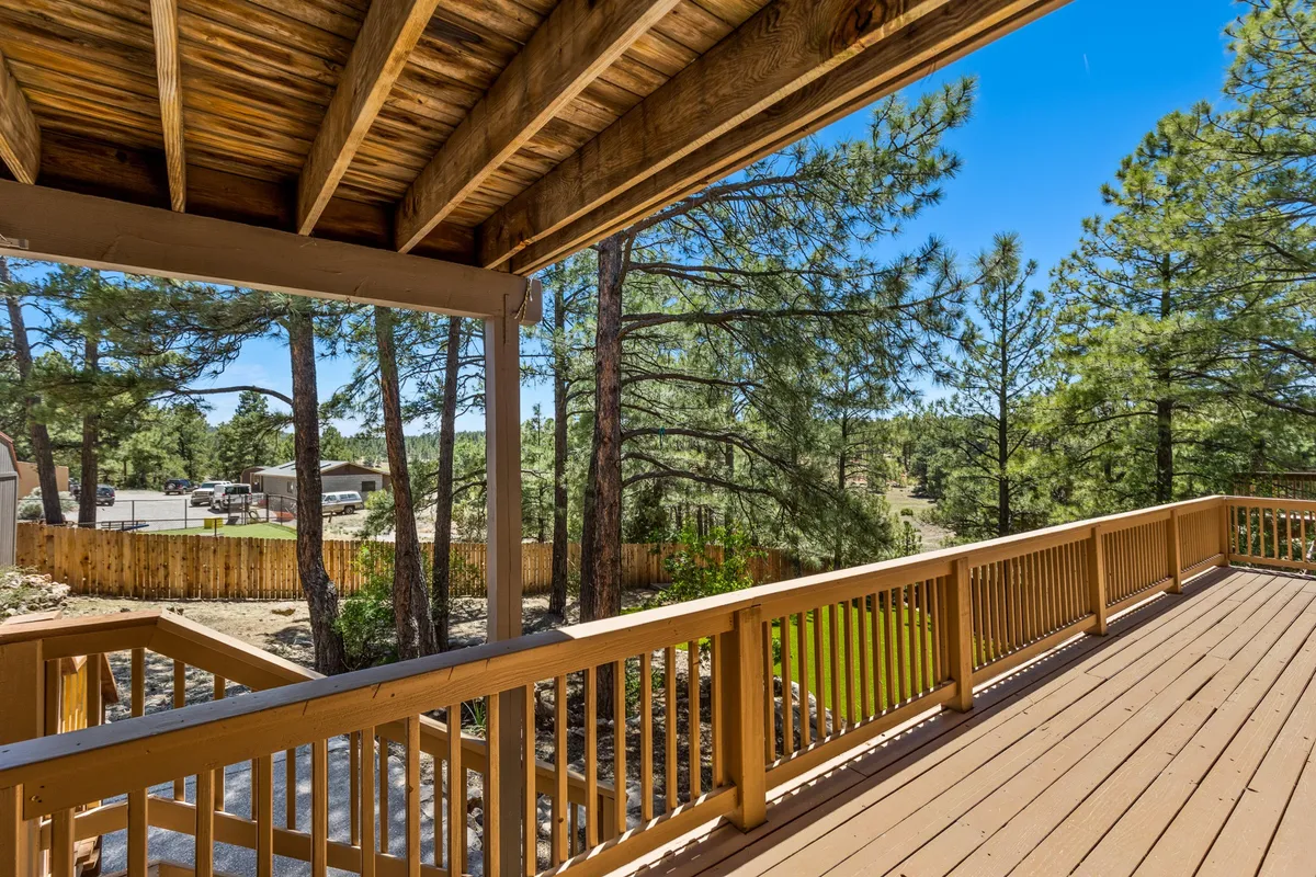 The image showcases a peaceful wooden deck with sturdy railings nestled among tall pine trees and set against a clear blue sky. The deck is inviting for relaxation and perfectly positioned for enjoying the natural surroundings just outside the home.
