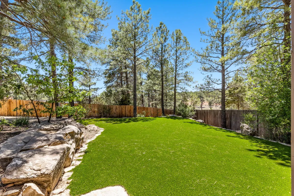 A lush green lawn bordered by tall pine trees, natural rock formations, and a rustic wooden fence. The open area is ideal for gatherings, outdoor games, picnics, and relaxation, set in a tranquil and inviting environment.