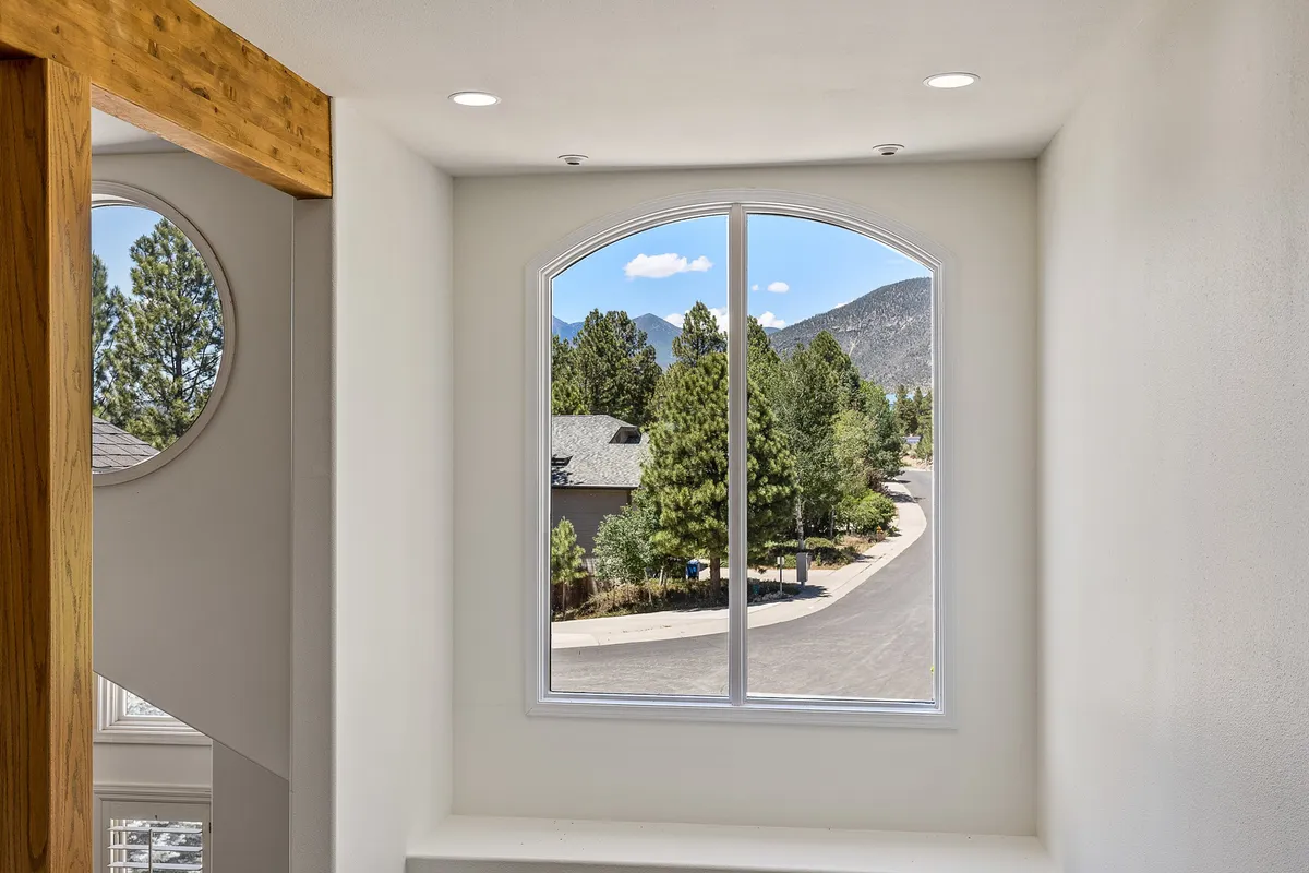A serene interior perspective with a large arched window offering expansive mountain views and abundant natural light, surrounded by trees. The inviting space is perfect for reading or simply soaking in the scenery after a day outdoors.