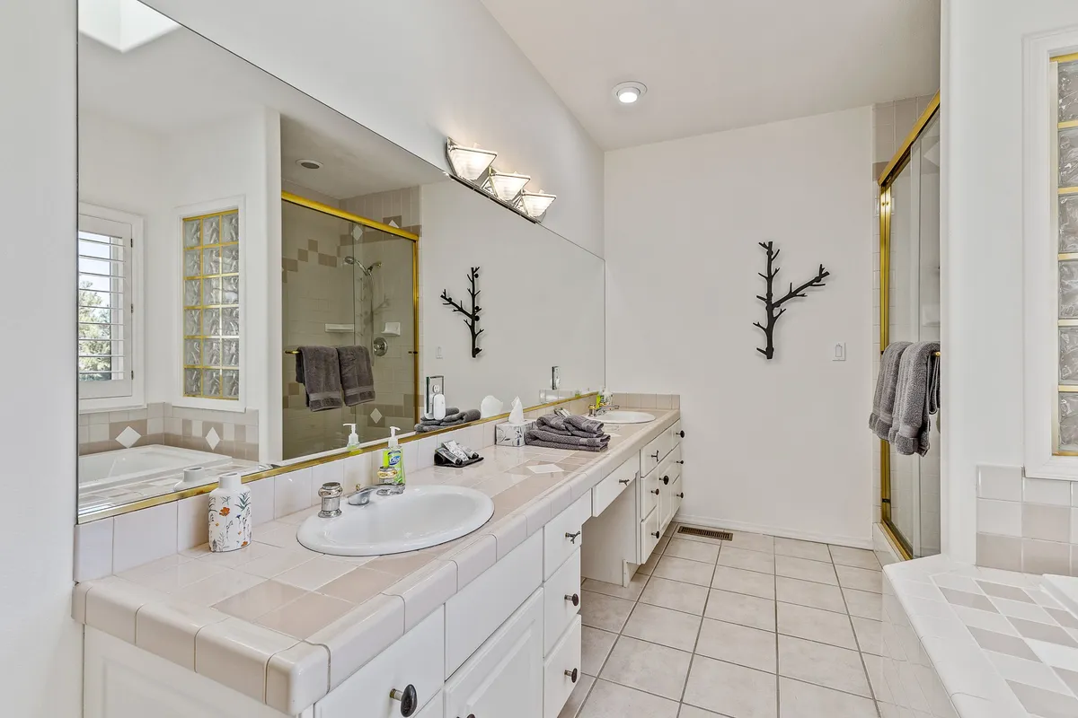 A well-appointed modern bathroom with double vanity sinks, a large mirror, a shower area, and a decorative wall branch. The space is bright, clean, and organized, suggesting comfort and convenience.