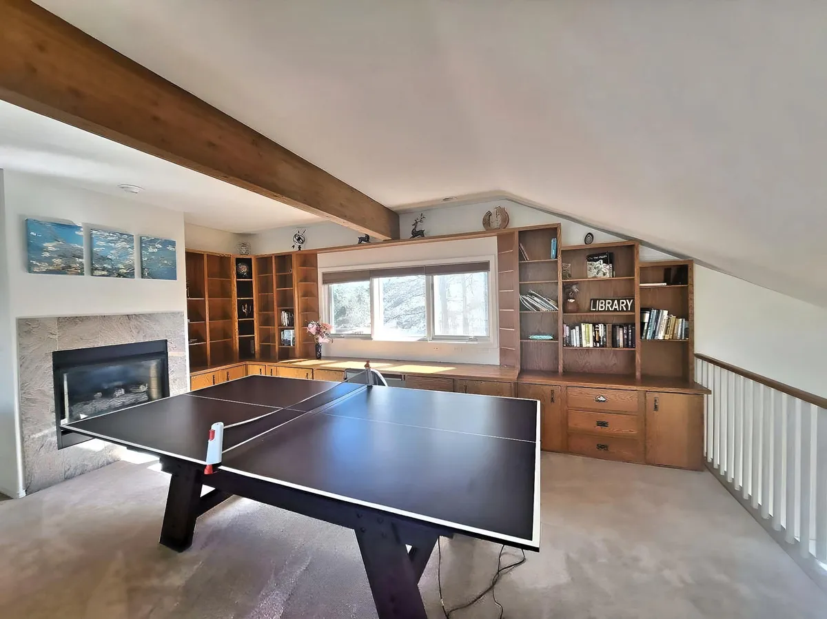 The image captures a cozy interior game room with a ping pong table as the focal point, surrounded by bookshelves, a fireplace, and a window that frames a view of the outdoors. The room exudes a relaxed, welcoming atmosphere ideal for friendly competition or unwinding with a book after a game.