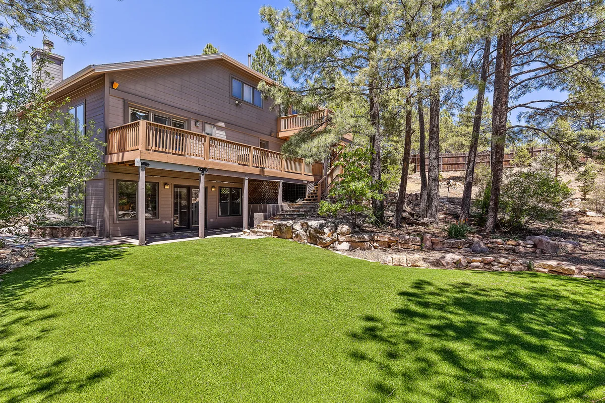 A spacious exterior deck opens onto a lush green lawn bordered by mature trees and stonework. The setup is perfect for outdoor gatherings, barbecues, or games like cornhole in the fresh mountain air. The scene feels lively yet inviting, suitable for both recreation and simple relaxation.