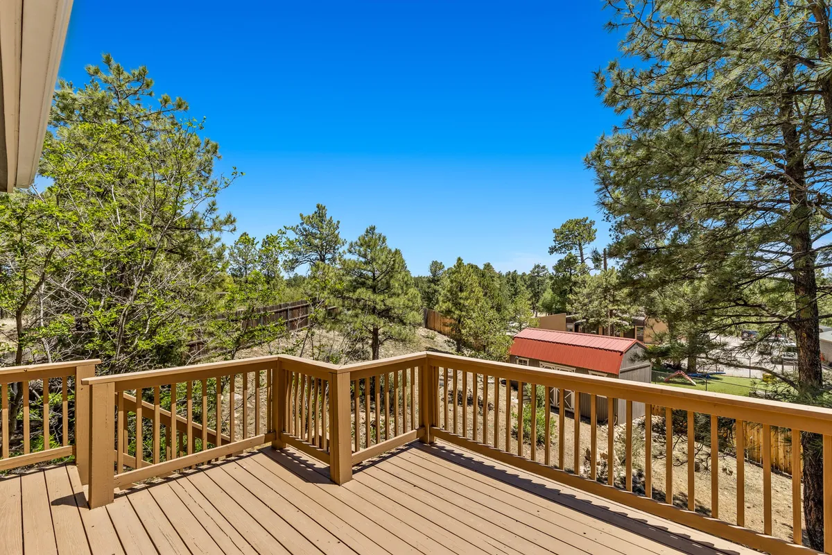 A wide wooden deck with a railing overlooks an expansive view of tall pine trees, with a distant red building adding a touch of urban color. The scene is tranquil, with the deck inviting for relaxation or social gatherings, and the natural setting hinting at Flagstaff’s unique blend of city and nature.